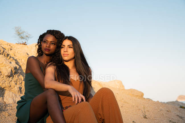 Мечтательные элегантные разнообразные подруги, сидящие на пляже — стоковое фото