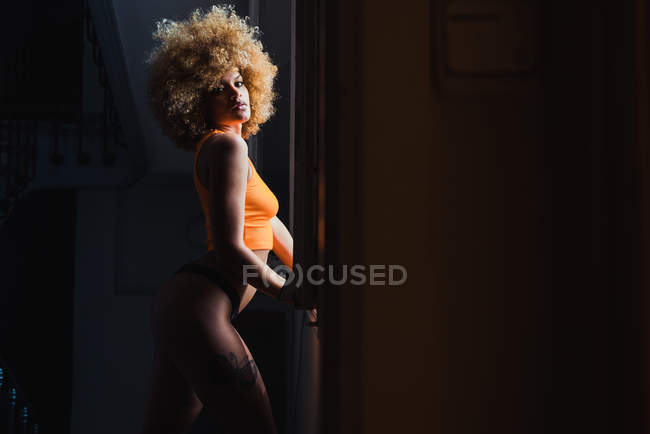 Alluring ethnic woman in lingerie standing in dark room — Stock Photo