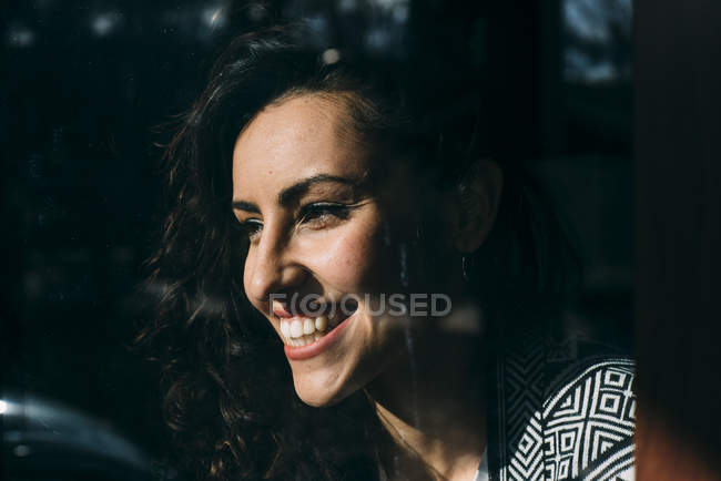 Sorrindo jovem mulher olhando pela janela sob a luz do sol — Fotografia de Stock