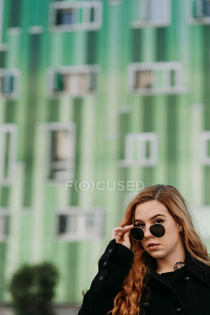 Élégante jeune rousse femme en lunettes de soleil regardant la caméra sur fond vert. — Photo de stock