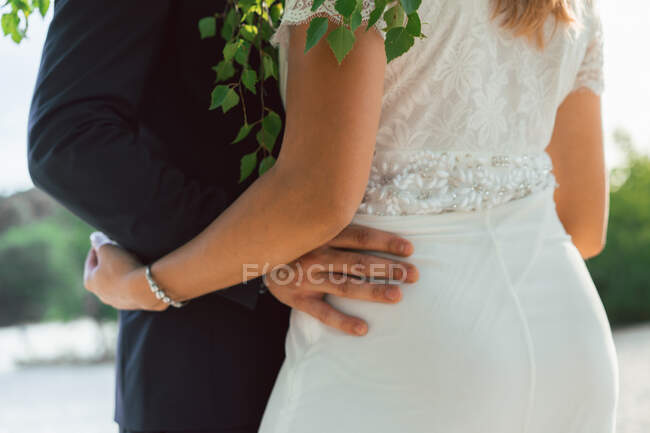 Faceless shot of groom embracing curvy bride in white dress standing under green tree in sunlight outdoors — Stock Photo