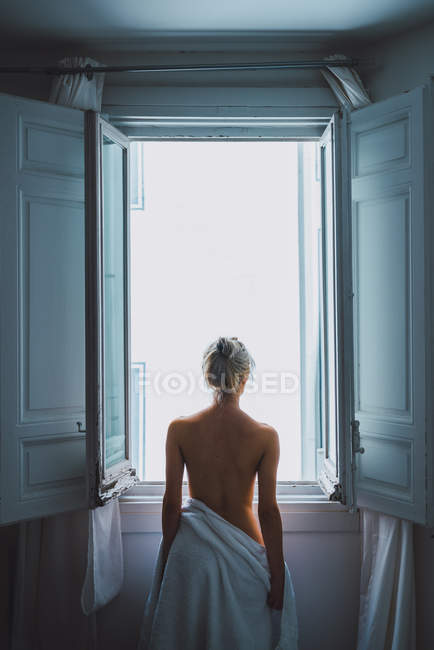 Vista trasera de mujer rubia desnuda con toalla blanca de pie cerca de la ventana abierta después de la ducha - foto de stock