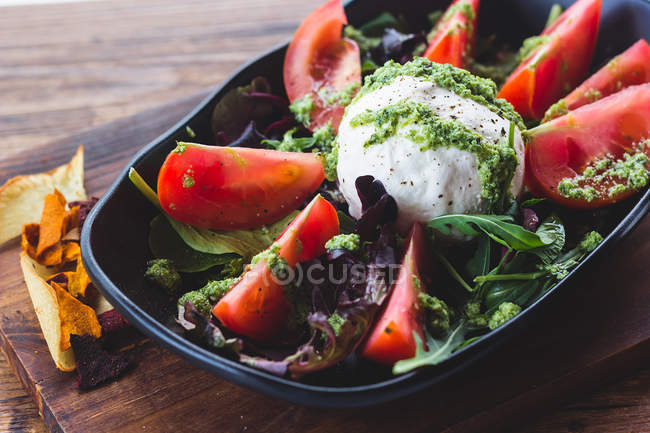 Close-up of served salad with tomatoes in green sauces on pile of greens with ball of cheese — Stock Photo