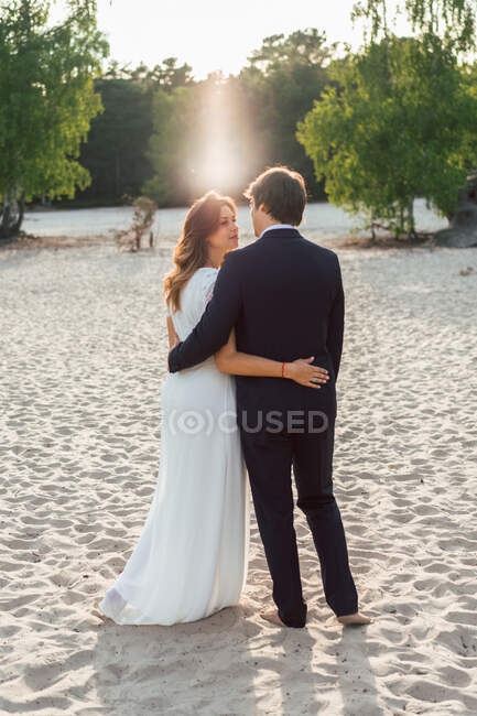 Liebender Mann umarmt schöne Braut in elegantem Kleid und schaut einander an, während sie an der Sandküste im Sonnenlicht steht — Stockfoto