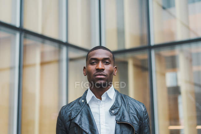 Confident ethnic man standing against glass building and looking at camera — Stock Photo