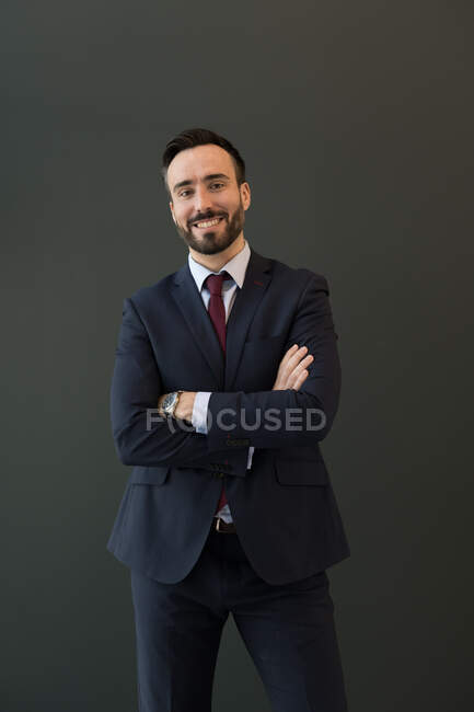 Smiling unshaven macho em trajes de negócios preto com relógio de pulso olhando para a câmera de pé com os braços dobrados sobre fundo preto — Fotografia de Stock