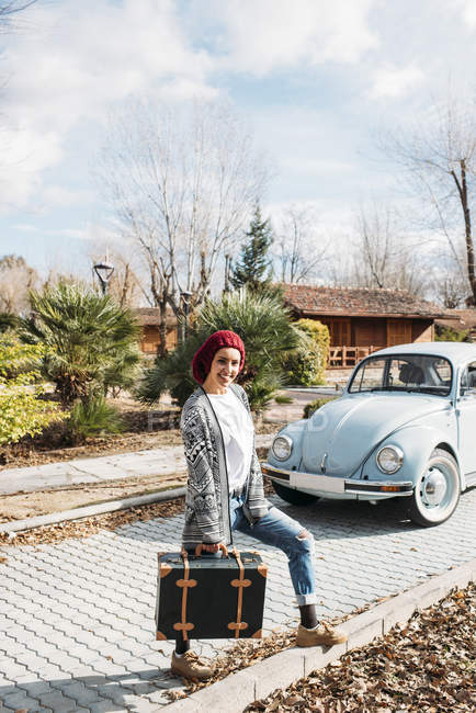 Молода жінка з старовинною валізою стоїть перед ретро-автомобілем і дивиться на камеру — стокове фото