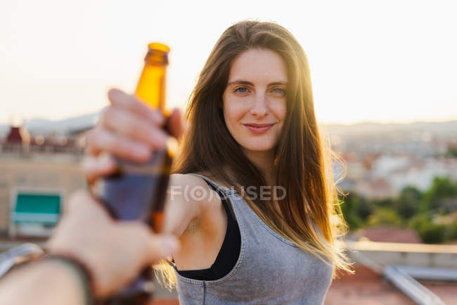 Mão humana dando garrafa de cerveja para a jovem mulher de pé no telhado e olhando para a câmera — Fotografia de Stock