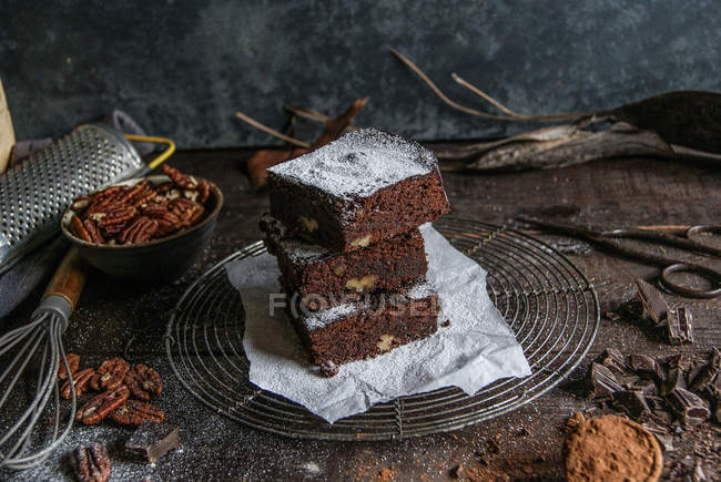 Trozos de delicioso chocolate brownie o n estante de alambre con ingredientes en la superficie de madera oscura - foto de stock