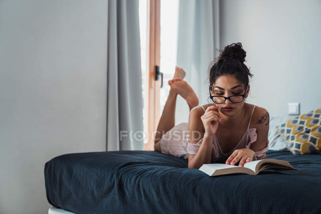 Focalizzata giovane donna lettura libro mentre sdraiato sul letto — Foto stock