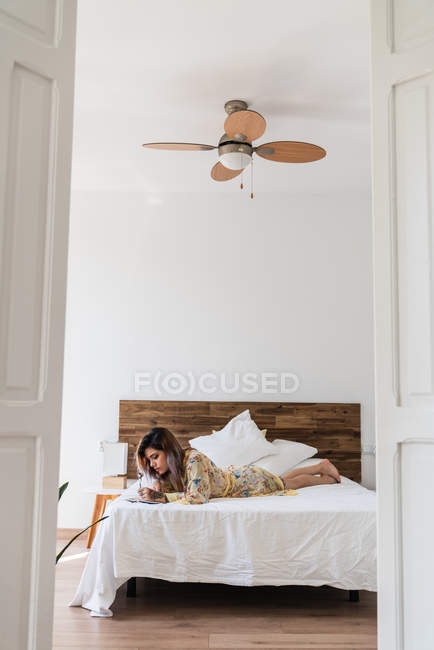 Jovem mulher em roupão de seda deitada na cama e fazendo esboços no bloco de notas no quarto elegante — Fotografia de Stock