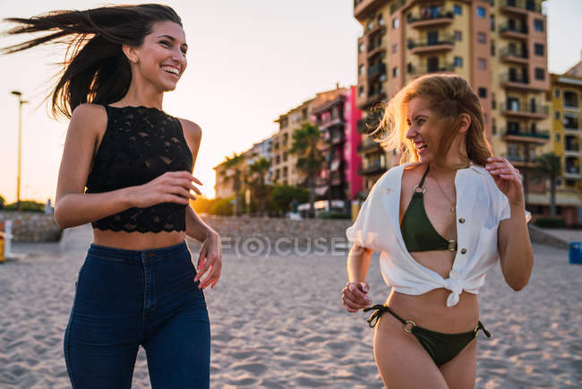 Смеющиеся подруги бегут по пляжу со зданиями на заднем плане — стоковое фото