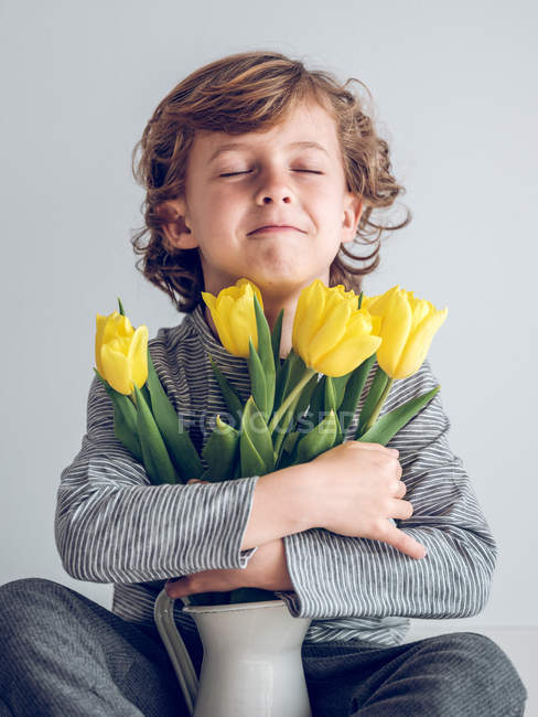 Young boy with eyes closed sitting and embracing bunch of yellow tulips on gray background — Stock Photo