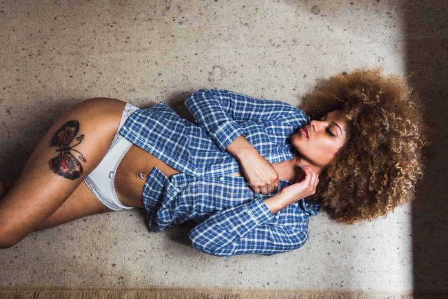 Joven mujer étnica en bragas y camisa a cuadros acostado en el suelo seductor - foto de stock