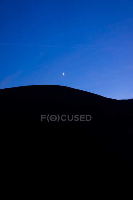 Мінімалістичний пейзаж чорного силуету гірських пагорбів на тлі сутінкового блакитного неба з півмісяцем — стокове фото
