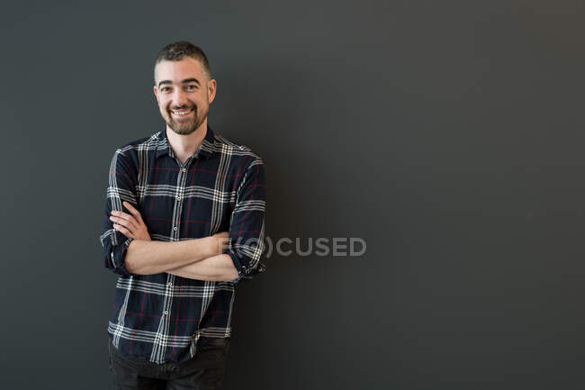 Handsome middle-aged bearded male with grey hair on temples in dark casual checkered shirt standing with crossed arms smiling and looking at camera over black background — Stock Photo