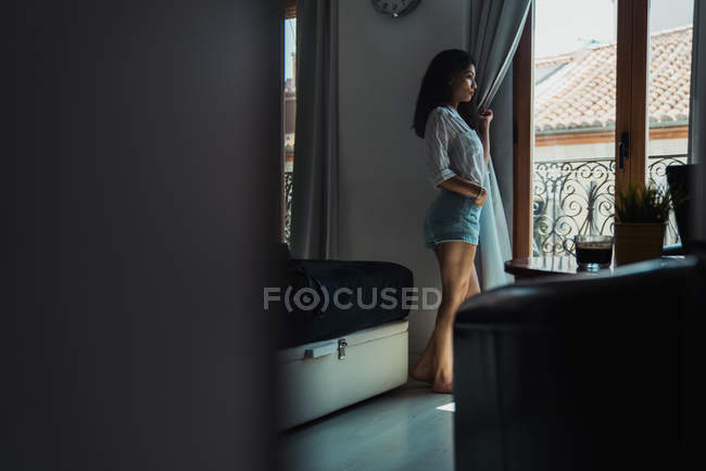 Dreamy woman in shirt and shorts standing near window — Stock Photo