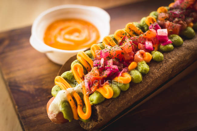 Close-up of delicious hot dog garnished with vegetables and sauce on wooden board — Stock Photo