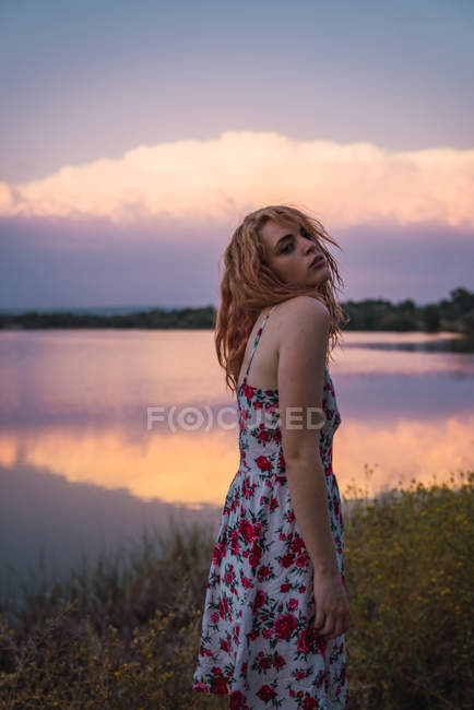 Young woman in summer dress standing on lake shore at sunset — Stock Photo