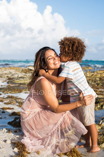 Adorable little boy standing on beach and kissing cheek of happily smiling mother at seaside in sunny day — Stock Photo