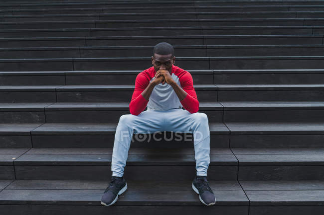 Etnia homem em sportswear sentado em cinza molhado escadas e apoiando a cabeça com as mãos dobradas e olhando para a câmera — Fotografia de Stock