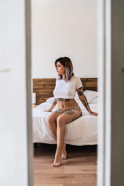 Mulher tatuada atraente em calcinha e camiseta sentada na cama e olhando para longe — Fotografia de Stock