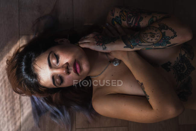 Topless tattooed woman covering breast and lying with eyes closed on floor — Stock Photo