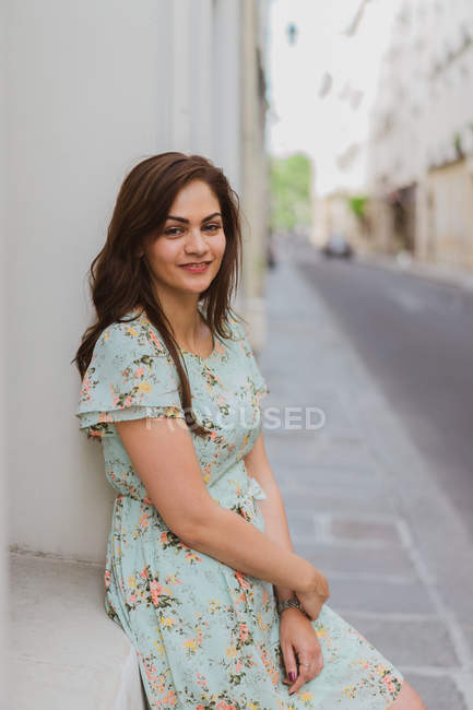 Smiling young woman in patterned summer dress leaning on wall on street and looking at camera — Stock Photo