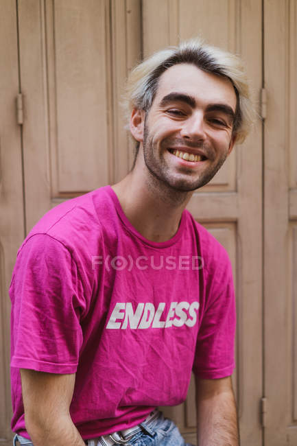 Homme blond souriant élégant assis contre une porte minable dans la rue et regardant la caméra — Photo de stock