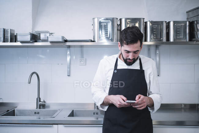 Vue latérale du jeune homme en uniforme de cuisinier appuyé sur le comptoir de la cuisine et utilisant un smartphone moderne tout en restant debout dans la cuisine du restaurant — Photo de stock