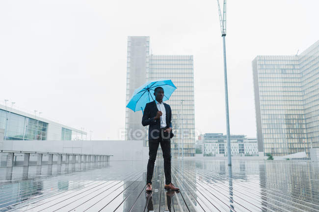 Empresario étnico sosteniendo paraguas azul mientras está de pie en pavimento húmedo en la ciudad - foto de stock