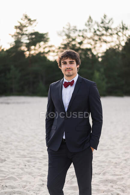 Uomo adulto che indossa elegante abito nero con papillon e in piedi sulla spiaggia con le mani in tasca guardando la fotocamera — Foto stock