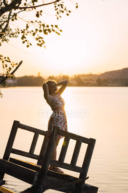 Frau im Kleid mit erhobenen Händen auf versunkenem Holzsteg am See bei Sonnenuntergang — Stockfoto