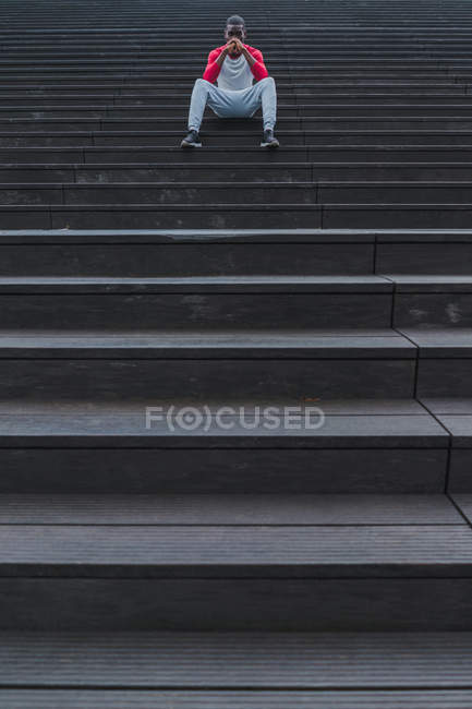 Ethnic man in sportswear sitting on grey wet stairs and propping head with folded hands and looking at camera — Stock Photo
