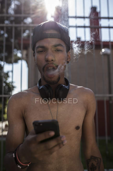 Afro young boy listening to music with headphones while smoking in front of grating — Stock Photo
