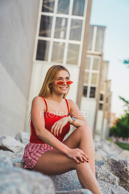 Mujer joven rubia en pantalones cortos y camiseta roja sentada sobre piedras en la ciudad - foto de stock
