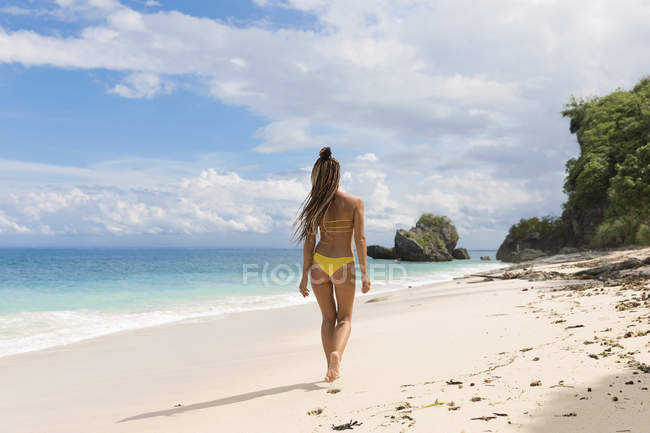 Visão traseira da mulher de biquíni amarelo andando na praia de areia no oceano — Fotografia de Stock