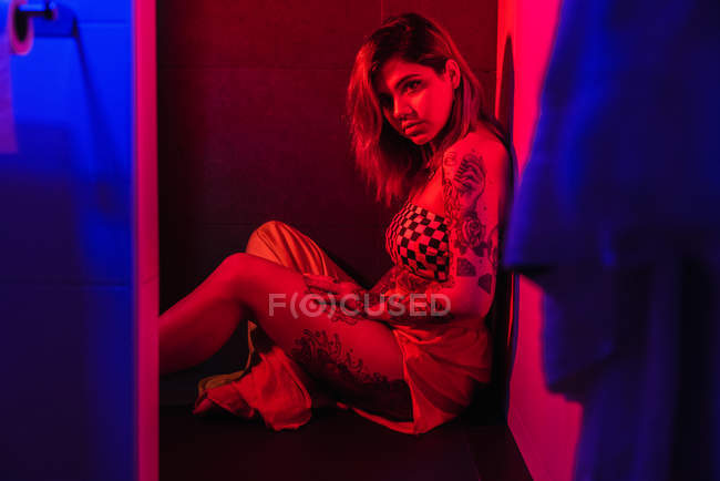 Jovem atraente no vestido sentado no chão no banheiro com iluminação vermelha brilhante — Fotografia de Stock