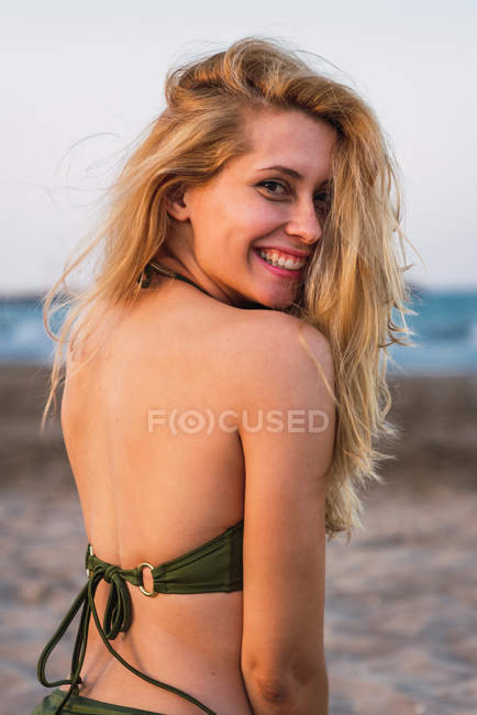 Cheerful woman in bikini sitting on beach and looking at camera — Stock Photo