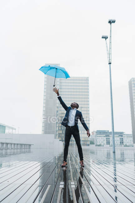Uomo d'affari etnico cattura ombrello mentre in piedi sul marciapiede bagnato in città — Foto stock