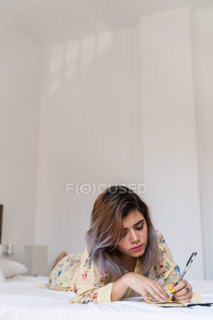 Mujer joven en bata de seda acostada en la cama y haciendo bocetos en bloc de notas en el elegante dormitorio - foto de stock