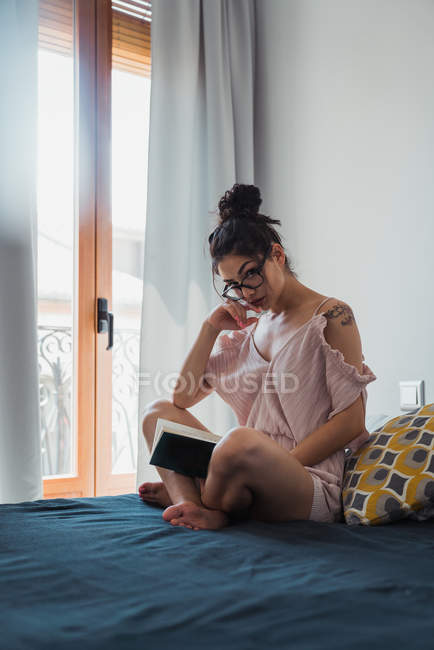Bruna donna con libro seduto sul letto e guardando la macchina fotografica — Foto stock