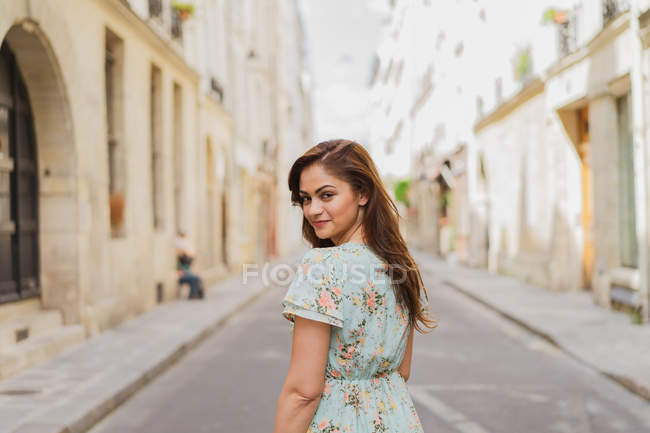 Усміхнена молода жінка в літній сукні ходить по вузькій вулиці і дивиться через плече — стокове фото