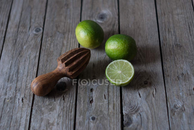 Limas frescas enteras y cortadas a la mitad con exprimidor de madera sobre madera gris - foto de stock