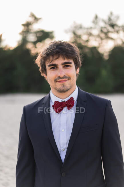 Adult man wearing elegant black suit with bow tie and standing on beach with hands in pockets looking at camera — Stock Photo