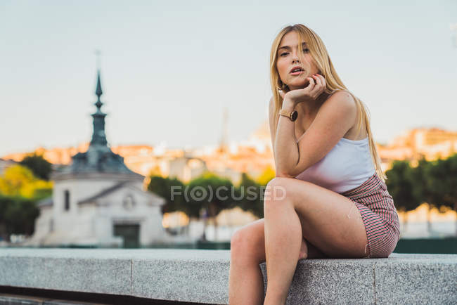 Junge Frau in lässigem Outfit blickt in die Kamera, während sie vor dem Hintergrund der Stadt sitzt — Stockfoto