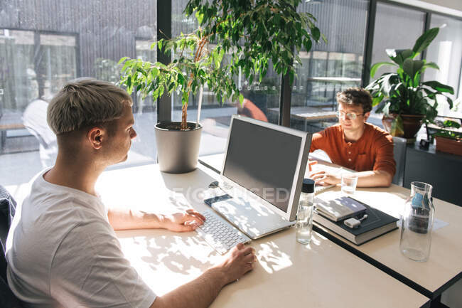 Männer sitzen im Großraumbüro an Tischen vor einander und arbeiten im Sonnenlicht an Computern — Stockfoto
