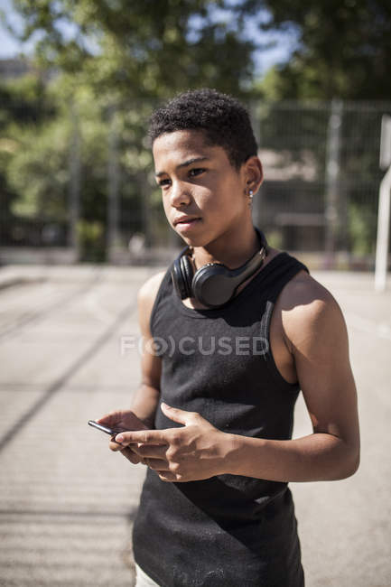 Afro jeune garçon écoute de la musique avec smartphone et écouteurs sur le terrain de basket — Photo de stock