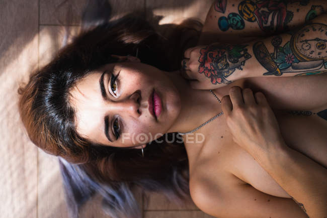 Encantadora mujer desnuda con tatuajes tocando collar y mirando a la cámara mientras está acostada en el suelo de madera - foto de stock