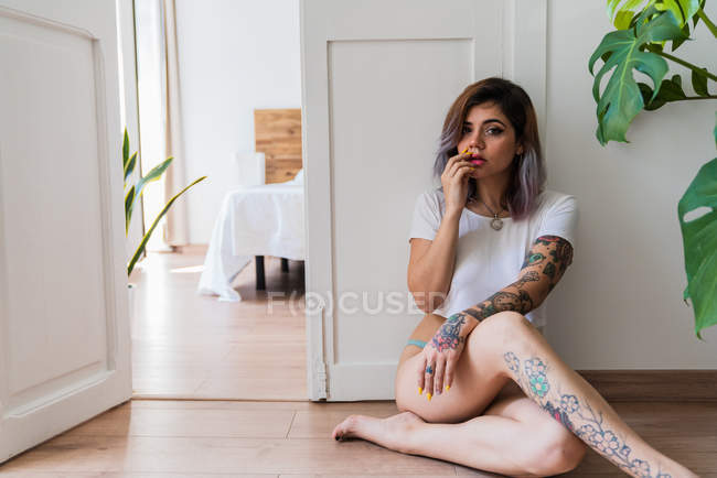 Barefoot woman with tattoos touching lips and looking at camera while sitting on floor near door of stylish bedroom — Stock Photo
